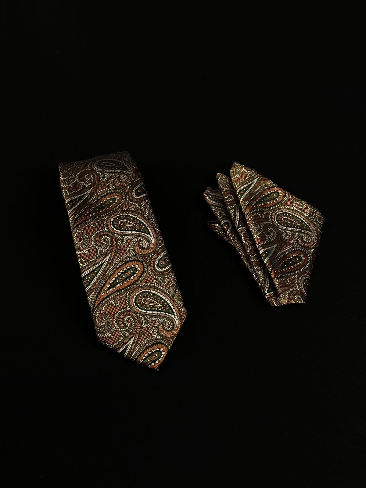 A matching necktie and pocket square set with an intricate paisley pattern in rust, olive and white.