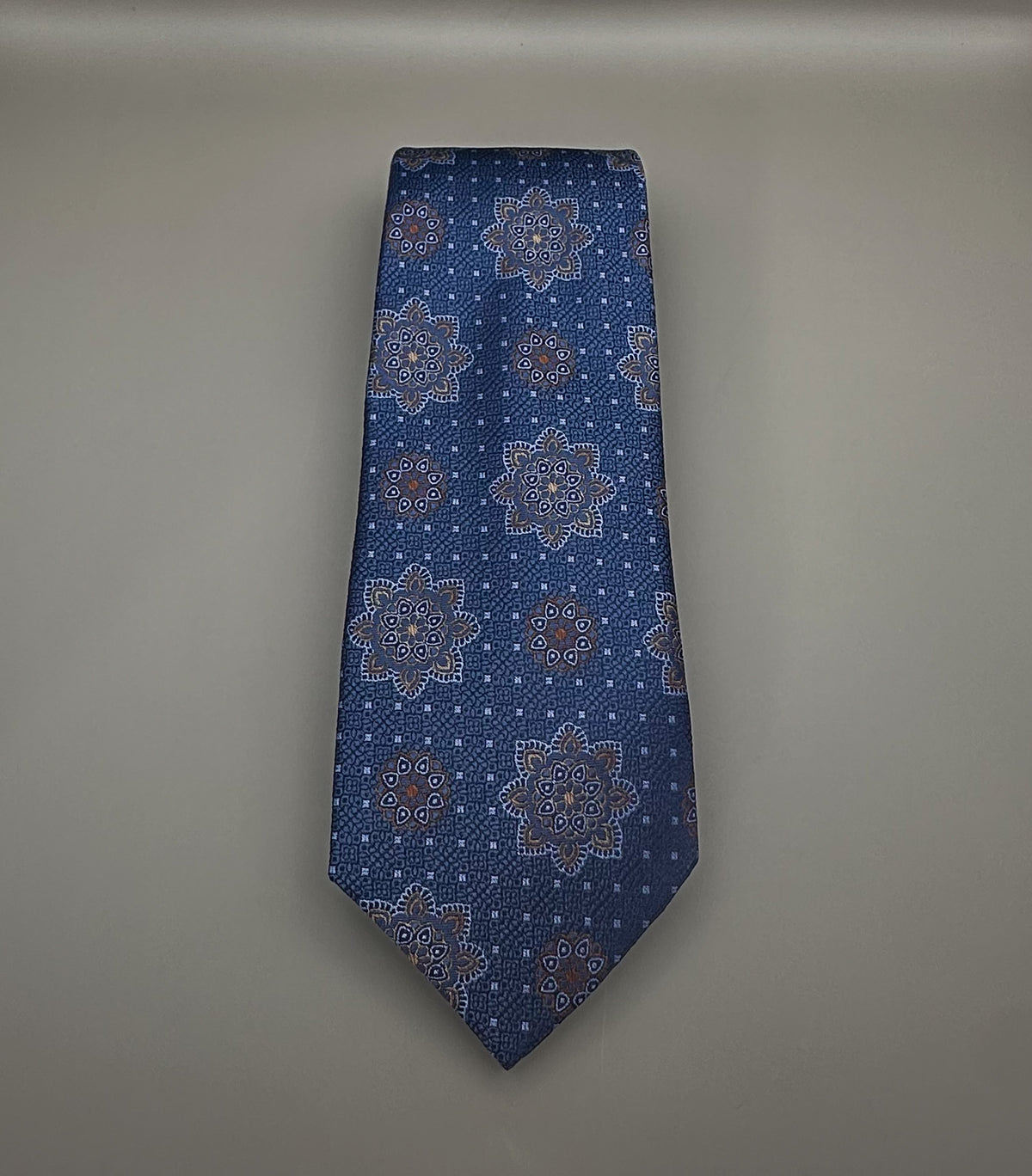 A classic necktie with a deep blue base and a detailed geometric medallion pattern in light blue, dark blue and tan.