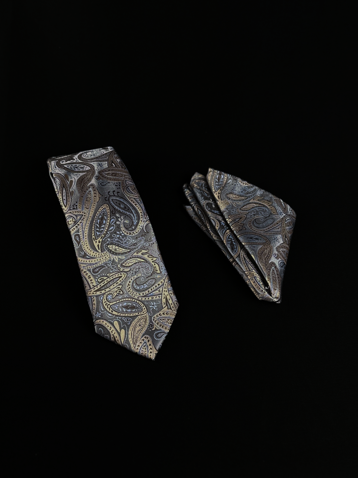 A matching necktie and pocket square set with an intricate paisley pattern in gray, gold, and beige.