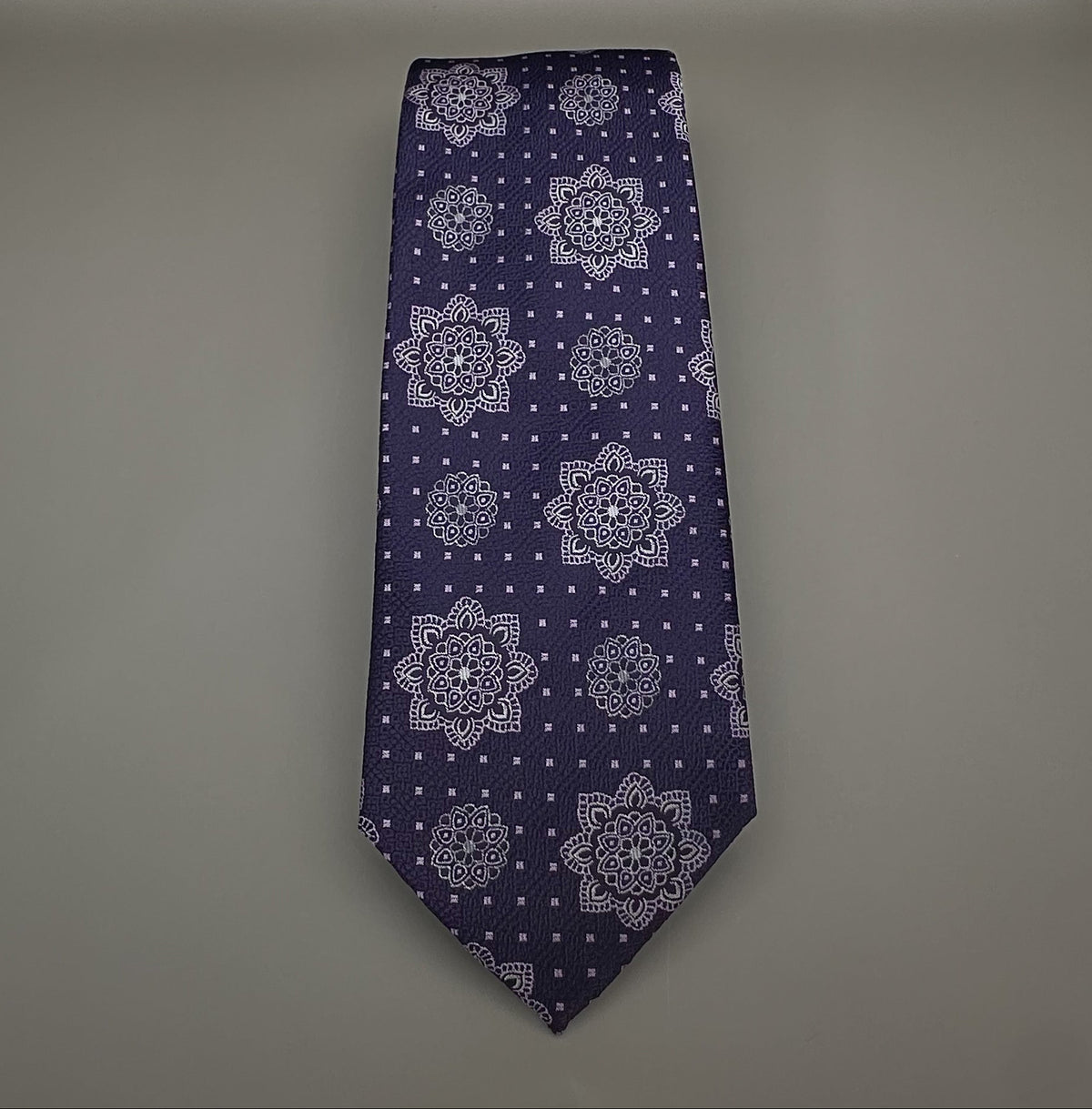 A classic necktie with a deep purple base and a detailed geometric medallion pattern in lavender, royal purple and silver.